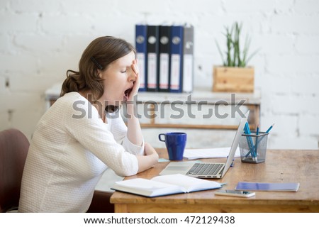 Portrait of young woman sitting at table in front of laptop, sleepy, tired, overworked, lazy to work. Attractive business woman yawning in home office relaxing or bored after work on laptop computer Royalty-Free Stock Photo #472129438