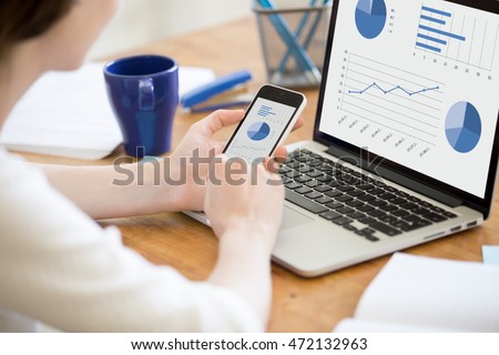 Close-up rear view of young business woman working in office interior on pc holding smartphone and looking at screen with diagrams. Office person using mobile phone and laptop Royalty-Free Stock Photo #472132963