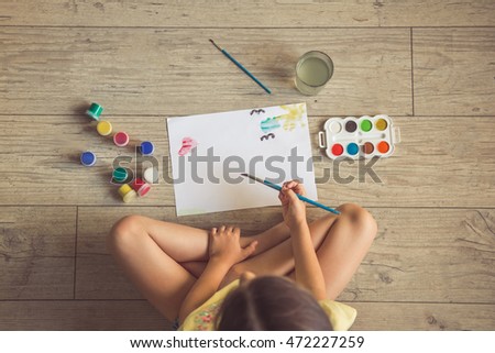 Top view of charming little girl painting using watercolors and gouache while sitting on the floor in her room at home