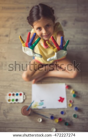 Top view of charming little girl showing her painted palms, looking at camera and smiling while lying on the floor in her room at home. Hands in focus