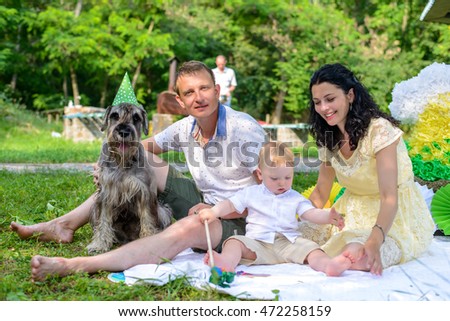 Young couple with a dog in fancy dress wearing a green party hat and photo booth glasses relaxing on a lawn in a lush park on a summer day