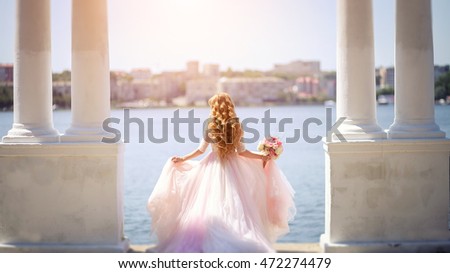 Girl standing in a beautiful long pink dress among the columns holding a bouquet of flowers, and turned his back