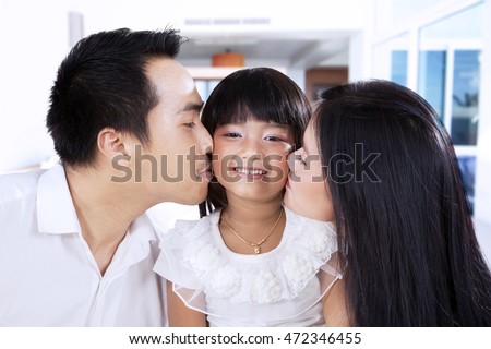 Photo of two Asian parents with black hair, kissing their daughter at home