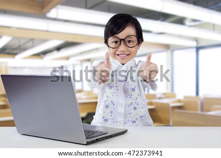 Portrait of happy little student showing OK sign with laptop computer on the table in the class