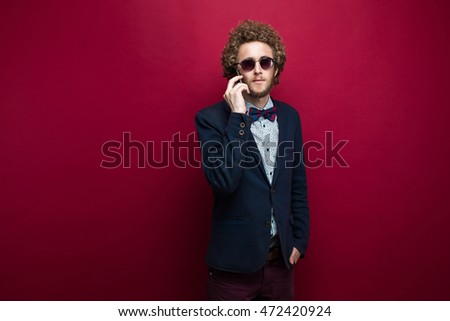 Elegant curly-haired man in suit and bow-tie and sunglasses using mobile phone with hand in pocket.Purple background.Isolate.