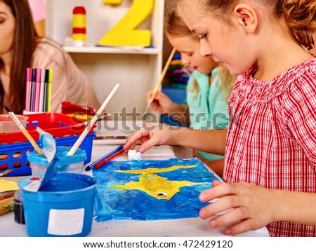 Group little girl and woman with brush painting on table in school. Painting box on foreground. Teacher help children draw pictures.