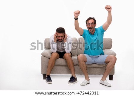 Picture of two men sitting on couch or sofa and looking at camera isolated on white background. Man in white shirt looking sad or disappointed. Emotions concept.
