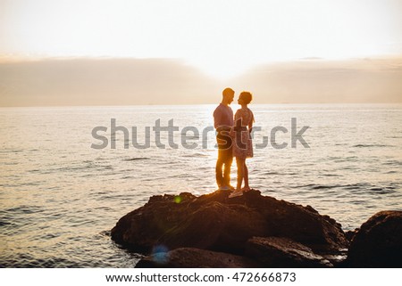 Bright evening sun shines over the couple posing on the stones by the calm sea Royalty-Free Stock Photo #472666873