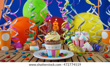 Birthday cupcake with candles burning on rustic wooden table with background of colorful balloons, plastic cups and candies with blue wall in the background                             