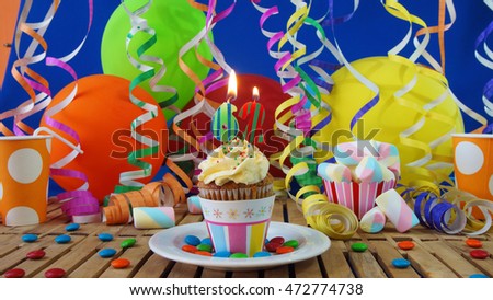 Birthday cupcake with candles burning on rustic wooden table with background of colorful balloons, plastic cups and candies with blue wall in the background                             