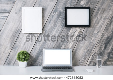 Creative designer workplace with blank white laptop computer, plant, mouse, water glass and two picture frames hanging above on wooden background. Mock up