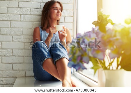 A well-pleased young girl enjoying an ideal day sitting in the window-sill. Wearing casual denim jeans and loose light-blue blouse, crossing legs and looking through the window Royalty-Free Stock Photo #473080843
