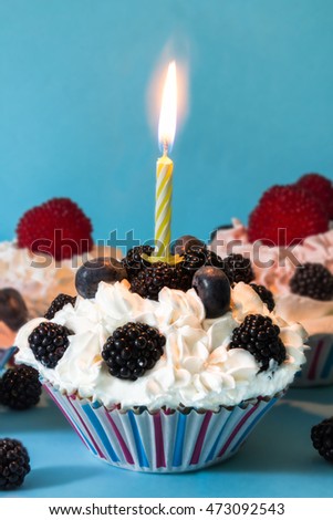 CUPCAKES WITH CREAM AND BERRIES, WITH A LIGHTED CANDLE ON BLUE BACKGROUND