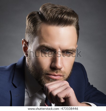 Young handsome bearded caucasian man with blue eyes sitting on chair. Perfect skin and hairstyle. Wearing blue suit. Studio portrait on gradient black to grey background.