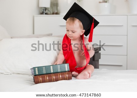 Funny portrait of 10 months baby boy in graduation cap looking at big books