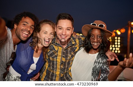 Multi-ethnic millennial group of friends taking selfie photo by mobile phone on rooftop terrace with a flash at night time
