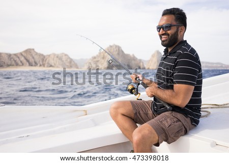 Young East Indian man fishing on open water from the boat. Beautiful sunny day with the shoreline visible in the background. South Asian male. Royalty-Free Stock Photo #473398018