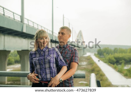 man and woman in the plaid shirt, trousers and chert, standing on the street holding hands against the background of the bridge. cloudy, rainy
