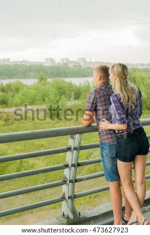 man and woman in the plaid shirt, trousers and chert, standing on the street holding hands against the background of the bridge. cloudy, rainy