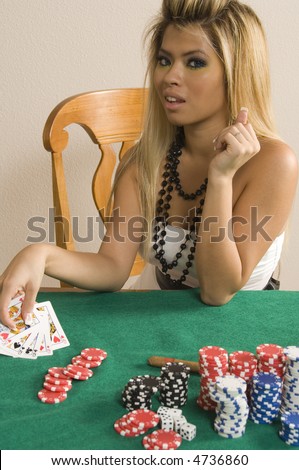 Pretty young Asian-American woman with blonde hair and black bead necklace holds poker hand by stacks of chips on green felt