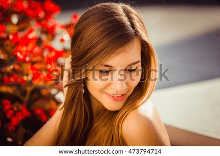 Stunning young woman leans her chin to the shoulder while sitting before red flowers