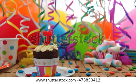 Birthday cupcake with candle burning on rustic wooden table with background of colorful balloons, plastic cups and candies with white wall in the background