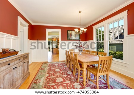 Red tones interior of American classic dining room. Long wooden dining table for ten person with decorative branches on the top, rustic cabinet and white wall trim. Northwest, USA