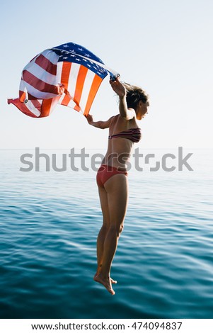 Back view of young woman in bright bikini in mid air above water surface with waving American flag against of sea blending with sky.