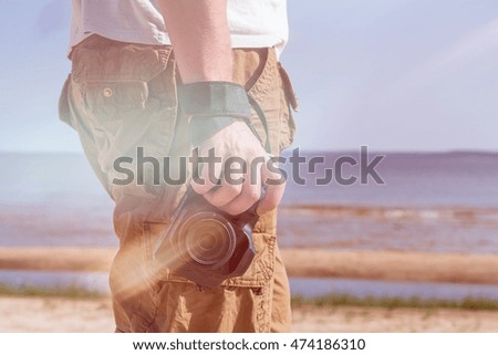 Man (male) dressed in shirt and breeches, standing on the sandy shore of the sea and holding a camera. Flare on photo.