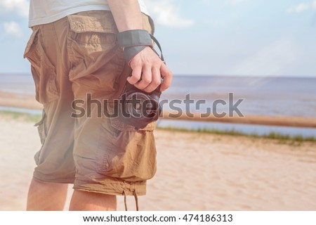 Man dressed in shirt and breeches, standing on the sandy shore of the sea and holding a camera. Flare on photo.