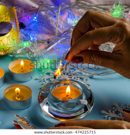 The girl's hands lit from the burning match of a candle. Dark red nail Polish. Blue background. Round candles. The twinkling lights of garlands. Carnival mask, decorative figurines of deer.