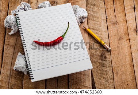 Blank diary with lined paper and red chili on top. With crumpled paper balls and yellow pencil on wooden table