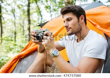 Concentrated young man tourist sitting in touristic tent and using old vintage camera in forest