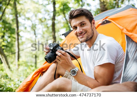 Cheerful young man tourist sitting and using photo camera in touristic tent in forest