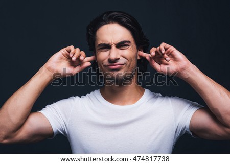 Too loud sound. Portrait of young man expressing negativity and covering ears by hands while standing against grey background