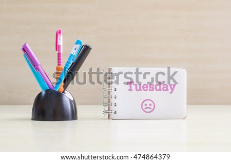 Closeup color pen with black ceramic desk tidy for pen and pink tuesday word in white page and frown face emotion at the note book on blurred wood desk and wood wall textured background 