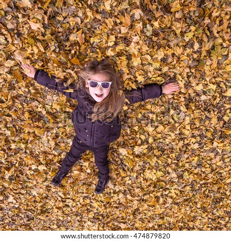 Little girl lying on a carpet of fallen autumn leaves in black jacket and sunglasses photoed from the top down