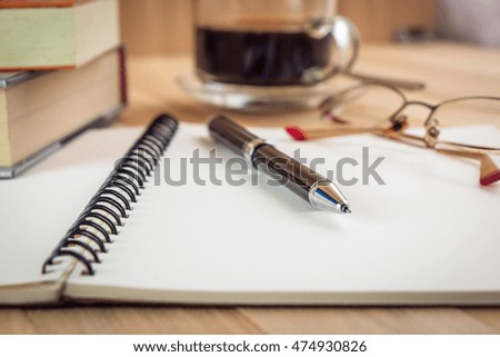 Put pen on blank notebook near glasses and Pile of thick book. Vintage style, Glass of coffee is blur background on wood table isolate.