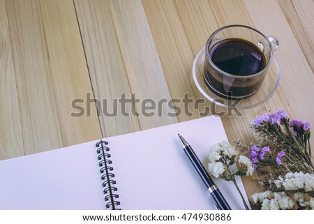 Put pen on blank notebook near the glass of hot and black coffee. Daisy and thin flowers are decorate on the wood table.