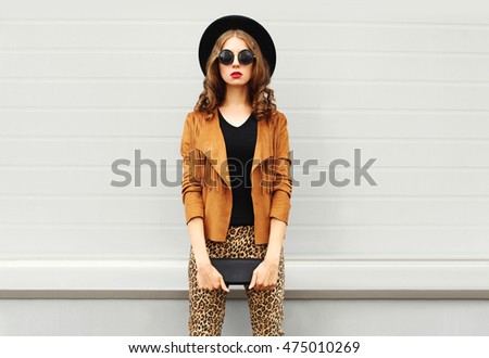 Fashion look, pretty woman wearing a retro elegant hat, sunglasses, brown jacket and black handbag clutch over grey background