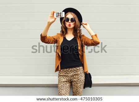Fashion pretty young woman model taking photo picture self-portrait on smartphone wearing retro elegant hat, sunglasses, brown jacket and handbag with curly hair over city grey background