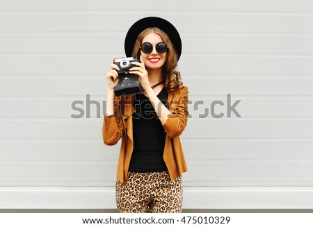 Fashion look, pretty cool young woman model with retro film camera wearing a elegant hat, brown jacket outdoors over city grey background