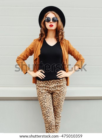 Fashion pretty woman wearing black hat, sunglasses and jacket over urban grey background