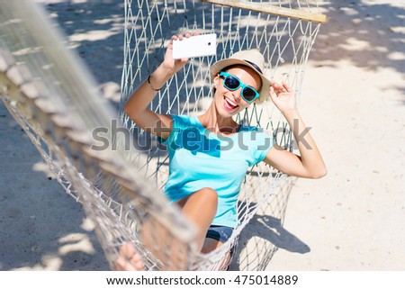 Vacation selfie. Enjoying the summer. Young pretty woman in hat and sunglasses using smartphone laying in hammock on the beach.