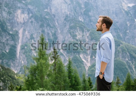 Young hipster man traveler hiking and watching the scenic landscape view. Travel lifestyle concept photo with beautiful mountain and evergreen trees on background. Summer vacations activity outdoor