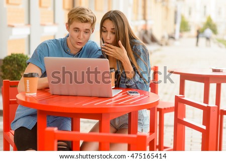 Surprised couple watches film online at the street cafe with the help of wi-fi connection. Girl touches her mouth with hand and sits with rounded eyes. Blond looks very excited.