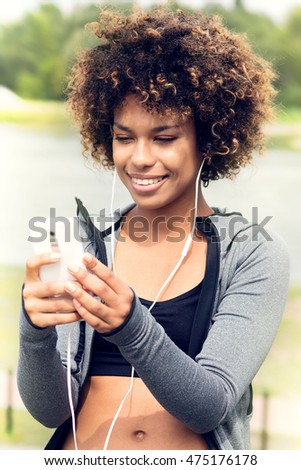 Photo of fit and sporty young woman listening to music with mobile phone. Beautiful african american girl with afro smiling. Outdoor shot.