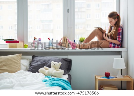 Thoughtful schoolgirl sitting on windowsill alone in her room, writing or drawing in notebook