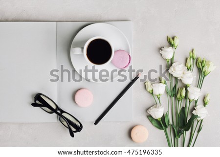 Coffee mug, empty notebook, pencil, glasses, white flowers and cake macaron on light table top view. Beautiful breakfast. Flat lay.
