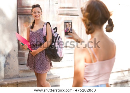 Smiling teenager student girl posing for a photograph on her first day at college entrance wooden door stone campus building, sunny exterior. Adolescents pupils using technology, outdoors.